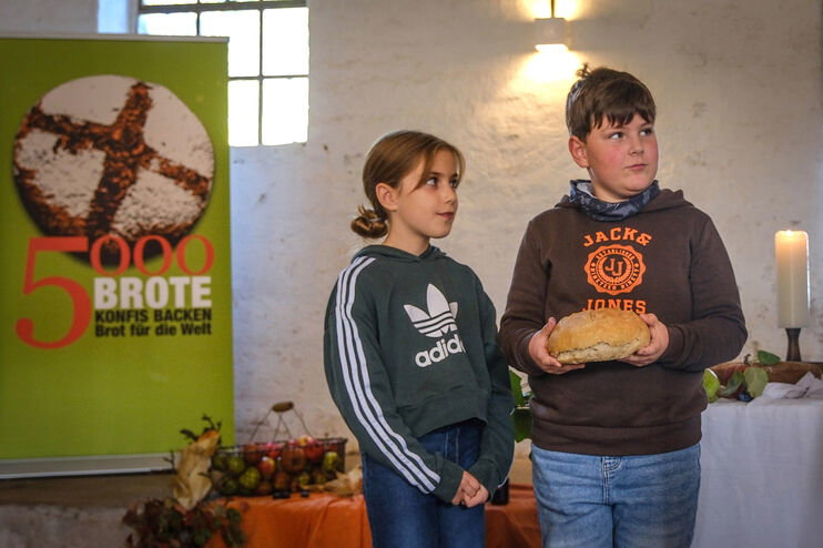 Eine als Mädchen und eine als Junge lesbare Person stehen mit einem Brot in einer Kirche neben einem Rollup mit der Aufschrift 5000 Brote.