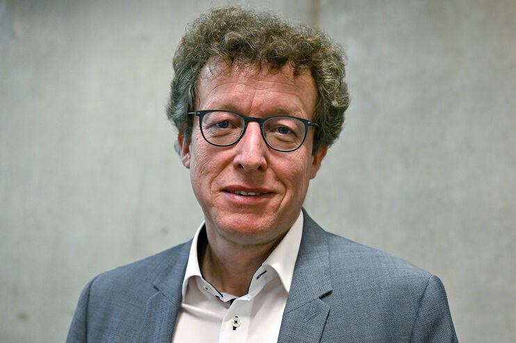 Eine als Mann lesbare Person mit Brille und Locken vor einer grauen Wand