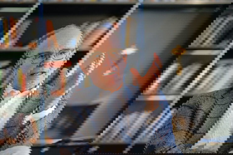 Eine männlich gelesene Person mit brauner Brillenfassung, kurzen grauen Haaren, grau-kariertem Jackett und hellblauem Hemd sitzt im Gespräch vor einem Bücherregal.