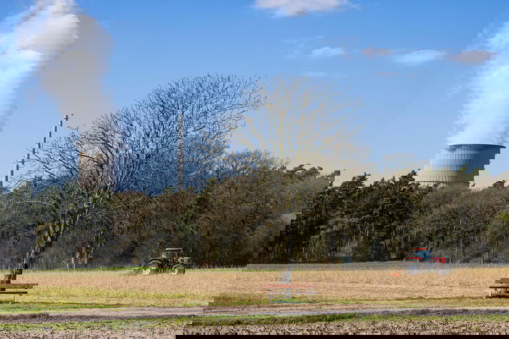 Eine Landschaft mit Baum und Bank; ein Trecker fährt über ein Feld, im Hintergrund quillt Dampf aus einem Kühlturm.