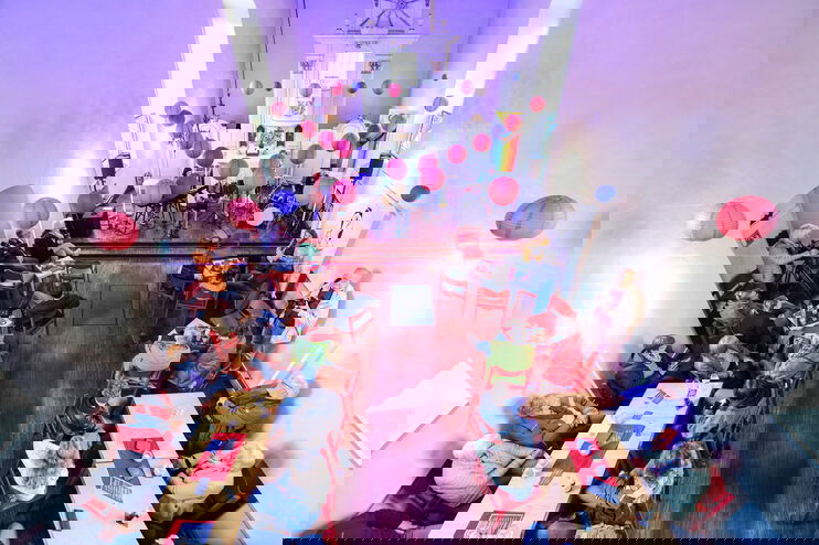 Blick von oben in einen Kirchenraum, in dem bunte Stühle an Tischen stehen, fast alle sind besetzt. Lila, rote und blaue Lampions sind quer durch den Raum gespannt.