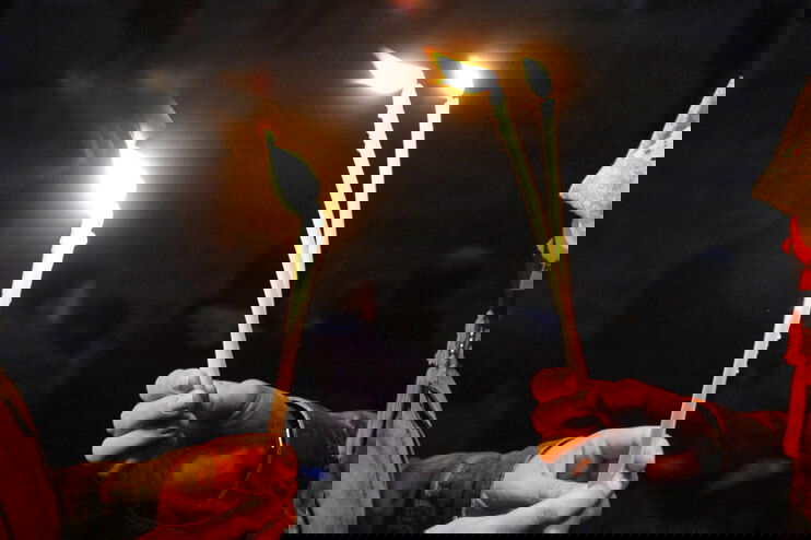 Zwei Menschen stehen sich gegenüber, zu sehen sind vor dunklem Hintergrund ihre Hände, die lange, dünne, brennende Kerzen halten.