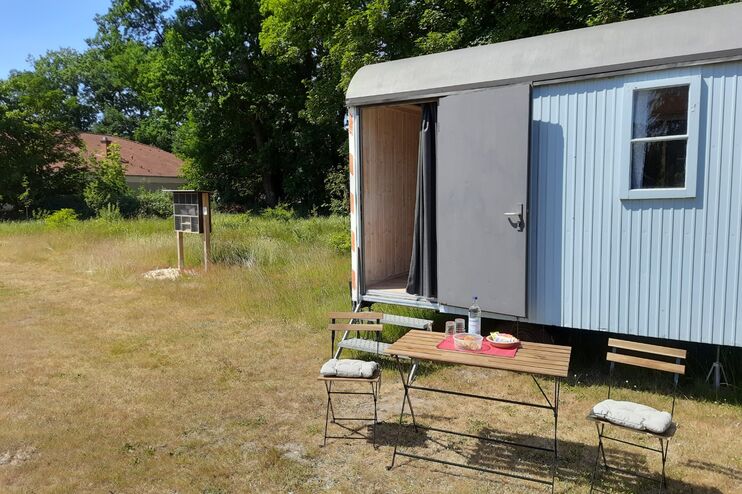 Auf dem Bild ist ein hellblauer Bauwagen auf einem Friedhof zu sehen. Seine Tür ist geöffnet. Vor dem Wagen stehen ein Tisch und zwei Stühle.