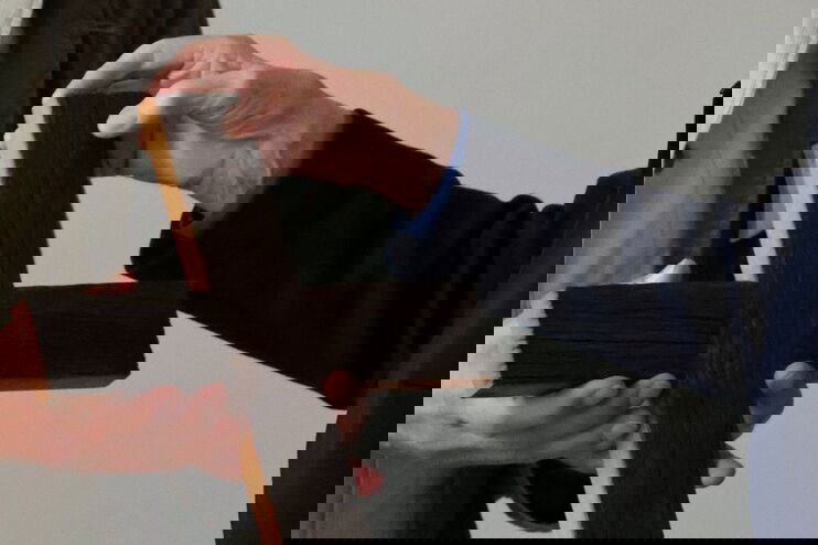 Eine Person übergibt einer anderen ein Holzkreuz. Sichtbar sind nur Hände und Schultern.