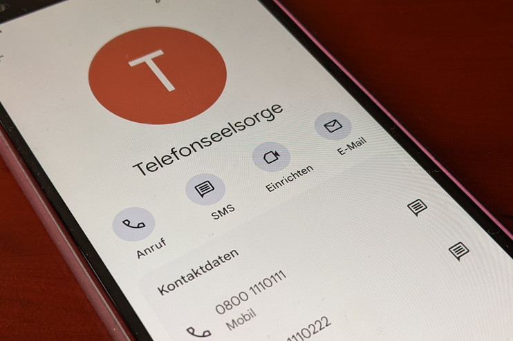 Ein Smartphone liegt auf einem dunklen Holztisch. Es zeigt den Kontakt "Telefonseelsorge" und die Nummern 0800 1110111 und 0800 1110222