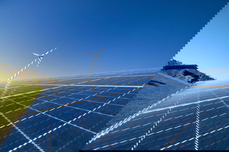 Eine große Photovoltaikanlage auf einer Freifläche, dahinter ein Windrad.