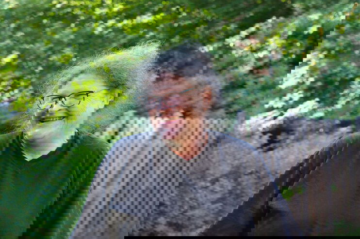 Eine Frau mit grauen Haaren, Brille und dunklem Shirt lacht, den Kopf nach rechts gedreht. Sie steht vor grünen Hecken und einem Holzzaun.