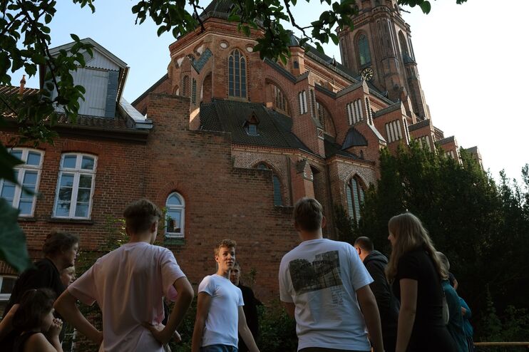 Mehrere junge Menschen stehen vor einer Kirche, sie sind von unten fotografiert. Die Sonne geht unter.