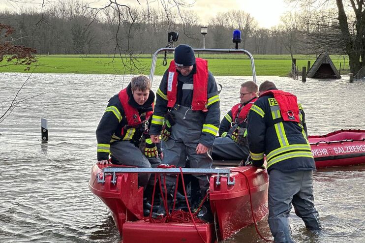Mehrere Personen mit Feuerwehrjacken sitzen in einem Boot, einer steht draußen im Wasser. Es hat eine Straße überflutet - ein Leitpfosten ragt nur noch ein Stück aus dem Wasser.