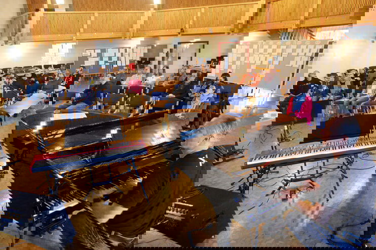 In einem großen Saal sitzt vorn rechts ein Mann am Flügel, auf Stühlen sitzt eine größerer Gruppe Menschen, die singt.