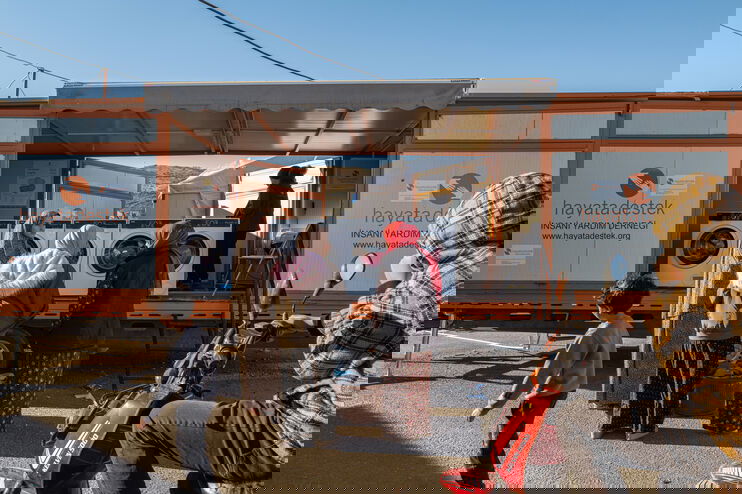 Auf einem Lkw, dessen Seite geöffnet ist, stehen Waschmaschinen. Davor stehen Menschen.