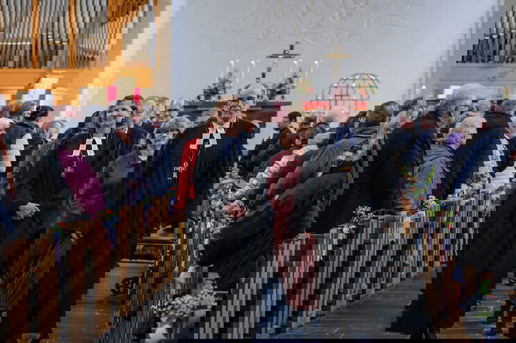 Zwei Personen in Talaren gehen zwischen mit Menschen gefüllten Bankreihen in einer Kirche hindurch. Im Hintergrund steht ein Kreuz an einer weißen Wand.