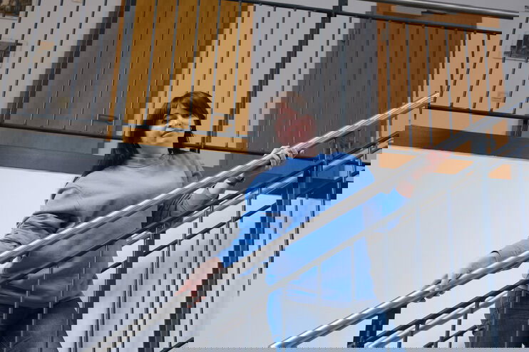 Eine als Frau lesbare Person mit dunklen Haaren und einem blauen Pullover steht an einem Treppengeländer.