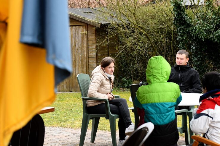 Fünf Menschen sitzen um einen Gartentisch auf einer Terrasse. Links, unscharf im Vordergrund, hängt eine blau-gelbe Fahne. 