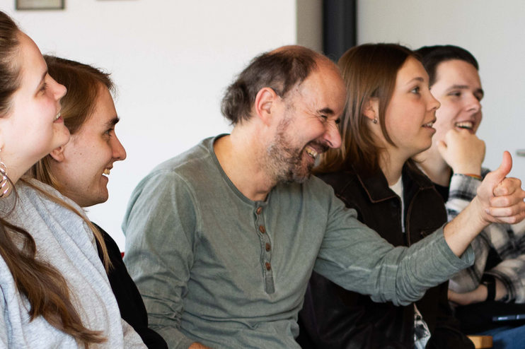 Eine männlich gelesene Person sitzt neben weiblich gelesenen, beugt sich leicht vor und zeigt den Daumen hoch. 