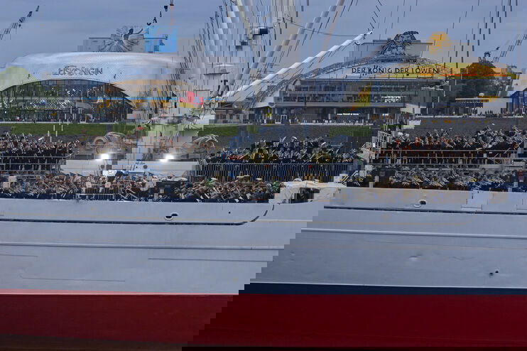 Blick auf die Seite eines Schiffs im Wasser. Das Deck ist sehr gut gefüllt mit Menschen, die teils Instrumente in Händen halten. 