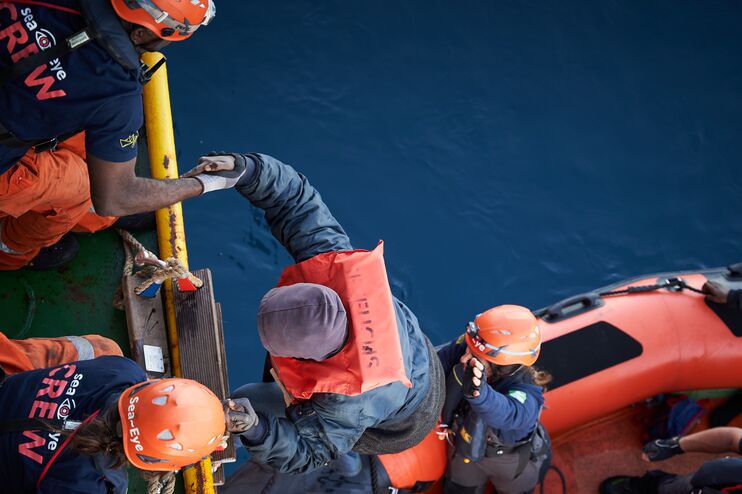 Blick von oben: Zwei Menschen auf einem Schiff helfen einem anderen aus einem Schlauchboot hinauf.