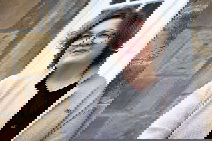 Eine als Frau lesbare Person lehnt an einer Hauswand.