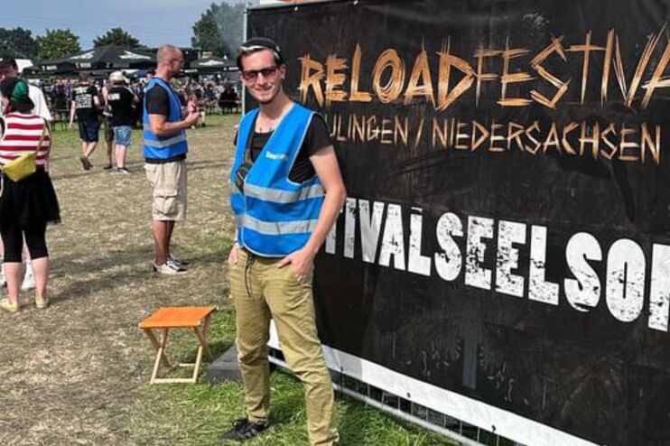 Ein junger Mann steht in einer blauen Wester vor einem Banner auf dem Festivalseelsorge steht.