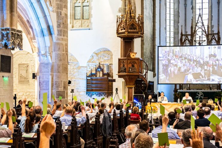 Eine Gruppe von Menschen in einer Kirche hält bunte Stimmzettel hoch.