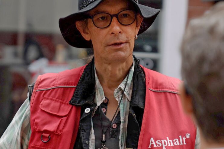 ein Mann in roter Weste verkauft einem Passanten eine Zeitung.