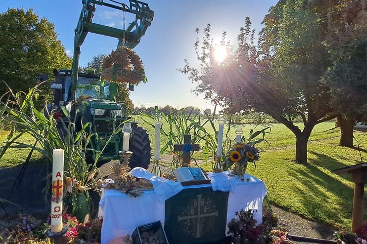 Ein Trecker steht hinter einem Tisch mit Gaben, einer weißen Tischdecke, einer Kerze als Altar. Dahinter sind ein Feld und Bäume erkennbar, die Sonne taucht alles in herbstliches Licht.