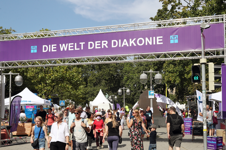 Auf einem großen lila Banner steht: Welt der Diakonie. Menschen in sommerlicher Kleidung gehen darunter hindurch.