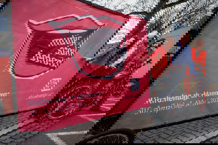 Zwei als Frauen lesbare Personen und eine als Mann lesbare Person halten ein Fahrrad, an das ein großes Kirchentags-Plakat angehängt ist.