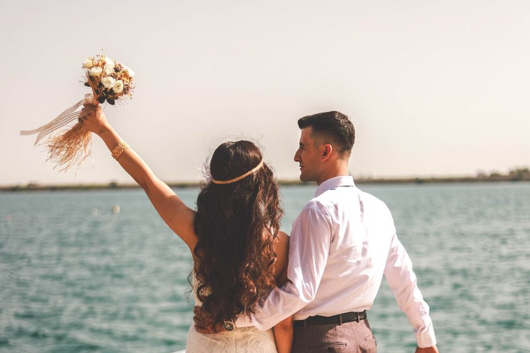 Eine weiblich und eine männlich gelesene Person in Hochzeitsoutfits auf einem Boot auf dem Wasser von hinten zu sehen. Die eine Person reckt einen Blumenstrauß in die Höhe.