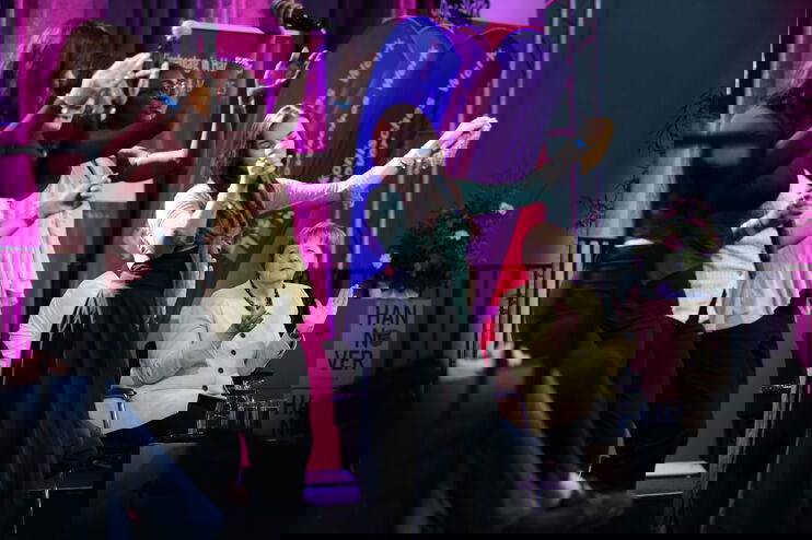 auf einer Bühne stehen mehrere weiblich lesbare Personen und singen, daneben sitzt Angela Merkel, eine weiblich gelesene Person, und klatscht in die Hände. 