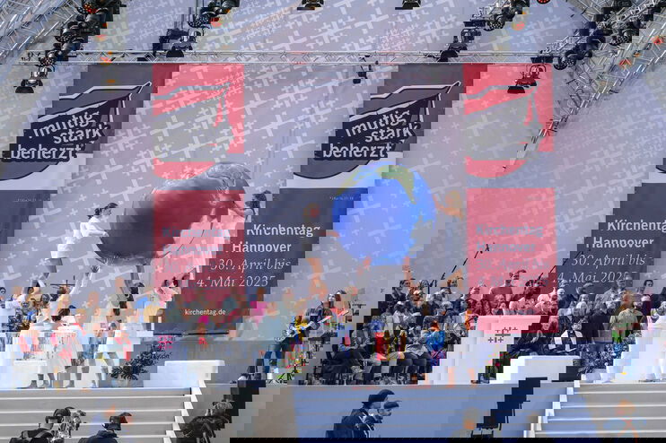 Ein Chor auf einer Bühne, davor mehrere Menschen in weiß, die einen riesigen Erdkugel-Ballon halten.