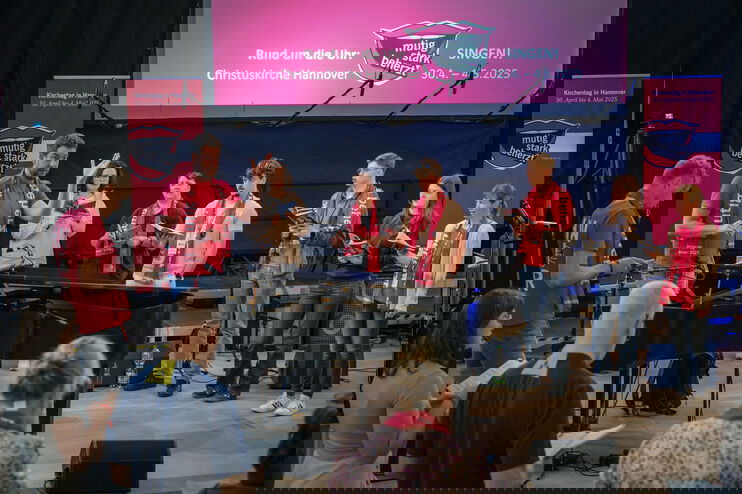 Eine als Mann lesbare Person steht hinter einem Flügel und dirigiert einen Chor.