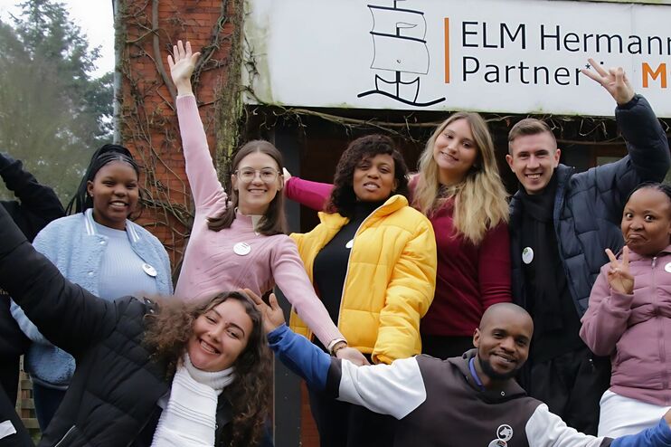 Ein Gruppe junger Menschen verschiedener Hautfarben steht vor einem Gebäude, auf dem steht ELM Hermannsburg.