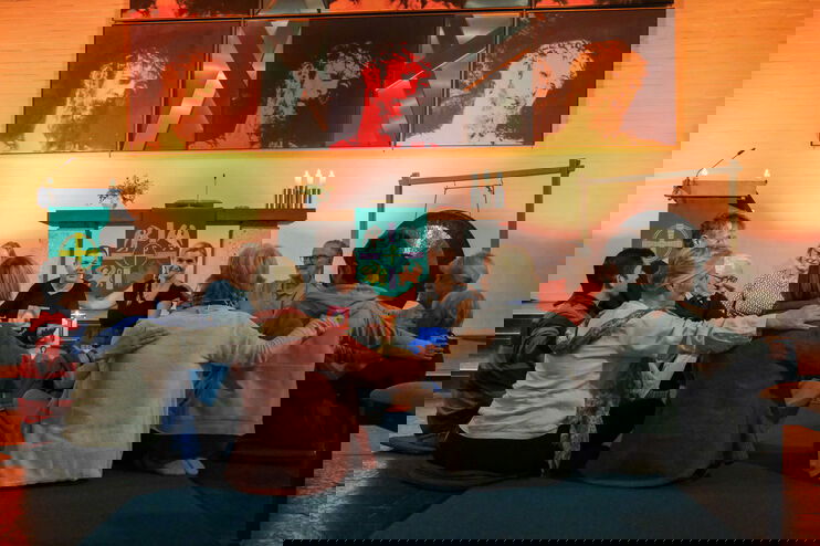 Eine Gruppe von Menschen sitzt im Kreis, jeder Person legt der rechts von ihr sitzenden die Hand auf die Schulter.