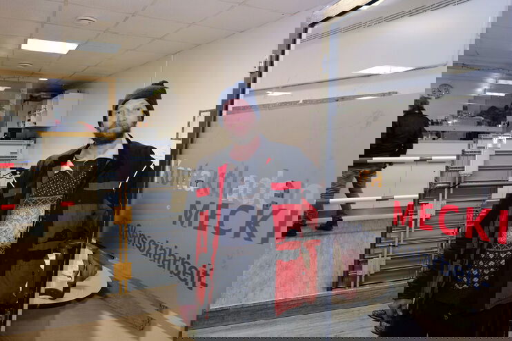 Eine als Mann lesbare Person mit Jacke und Mütze hält eine Tür mit Aufschrift Straßensozialarbeit auf.