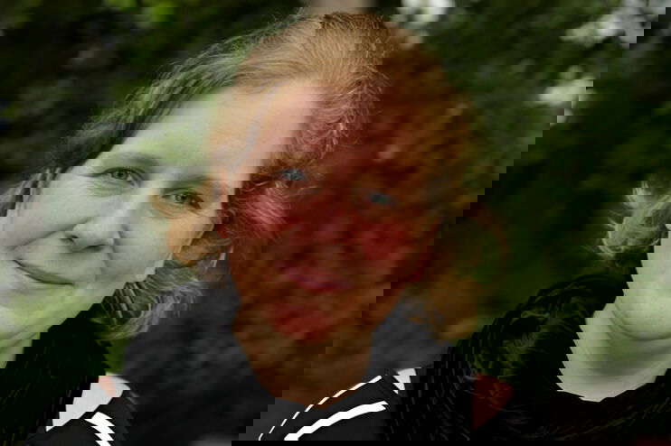 Eine junge als Frau lesbare Person vor einem Gebüsch im Portrait.