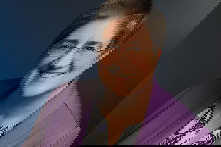 Eine weiblich gelesene Person mit Brille und grauen Haaren in Bluse und lila Jackett.