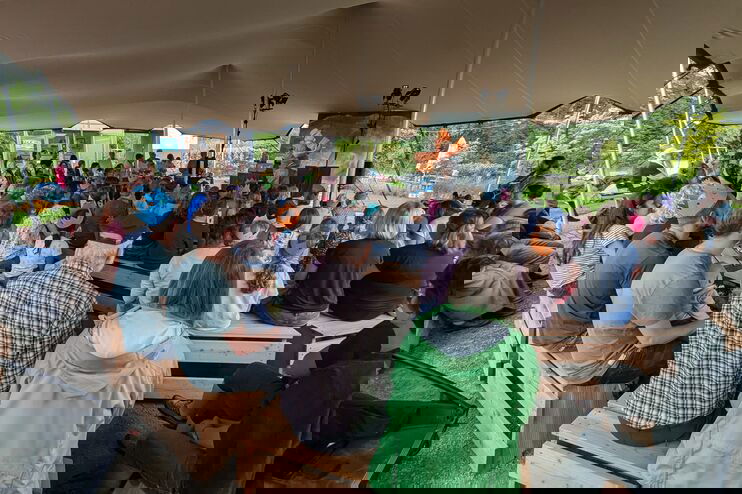 Viele Menschen sitzen unter einem Zelt im Grünen.