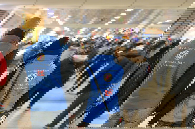 Zwei Personen mit blauen Westen, auf denen "Bahnhofsmission" steht, stehen im vollen hannoverschen Bahnhof.