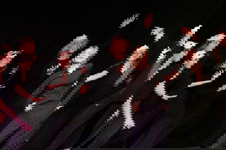 Eine Gruppe junger Menschen in schwarzer Kleidung auf einer Bühne, einige davon mit Maske.