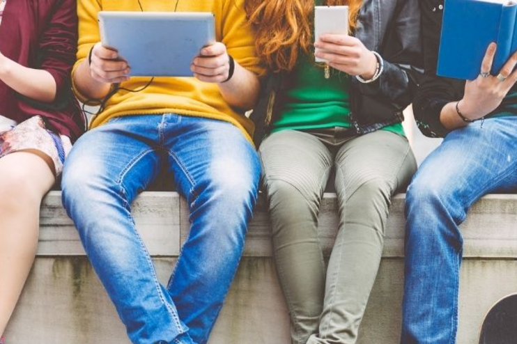 Vier junge Menschen sitzen auf einer Balustrade und haben Tablets, Handys und ein Buch in Händen.