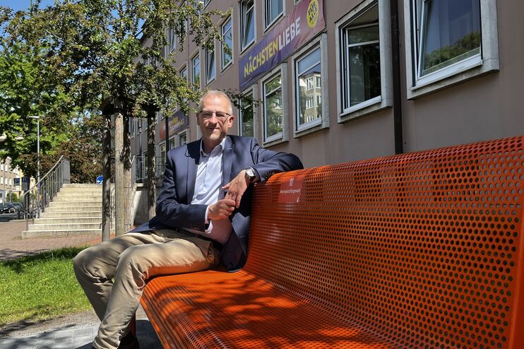 eine männlich gelesene Person sitzt auf einer orangefarbenen Bank draußen vor einem Gebäude