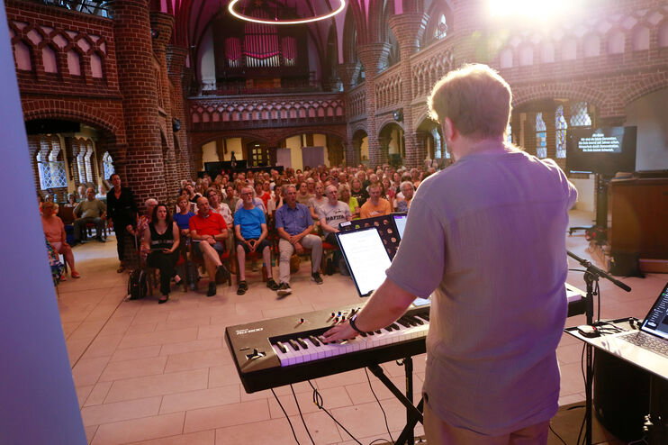 Eine als Mann lesbare Person steht am Keyboard und singt mit vielen Menschen in einer Kirche.