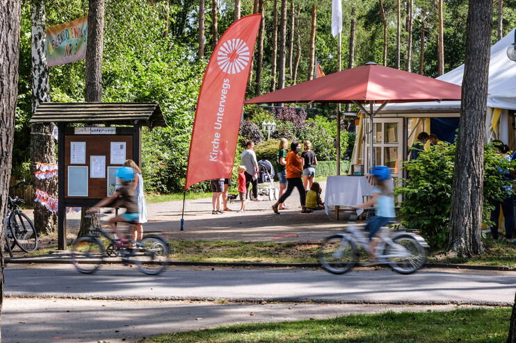 Eine Beachflag mit der Aufschrift „Kirche unterwegs“ auf einem Campingplatz. Im Vordergrund fahren Kinder auf einem Fahrrad.