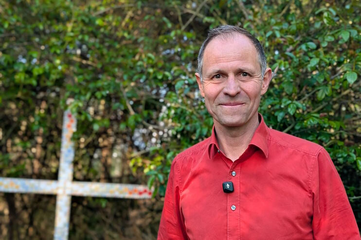 Eine als Mann lesbare Person mit rotem Hemd vor grünen Sträuchern, neben ihm ein angemaltes Holzkreuz.