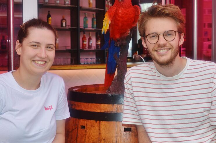 Links ist eine Frau und rechts ein Mann vor einer Bar im Hintergrund.