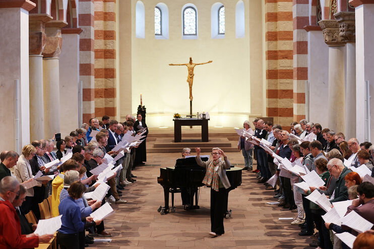Viele Menschen singen in einer Kirche. Eine als Frau lesbare Person dirigiert, eine andere spielt am Flügel.