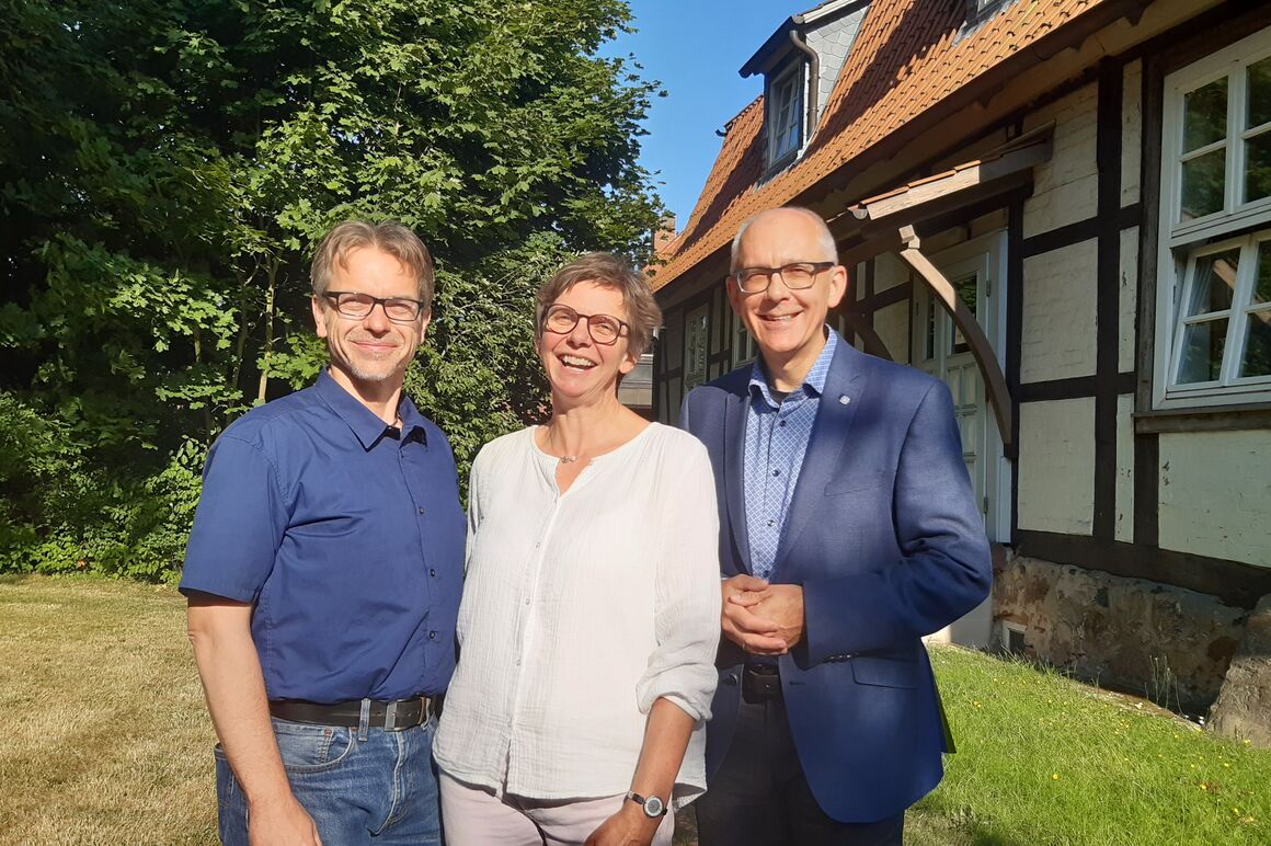 Zwei männlich und eine weiblich gelesene Person stehen vor einem Fachwerkhaus zusammen und lächeln in die Kamera.