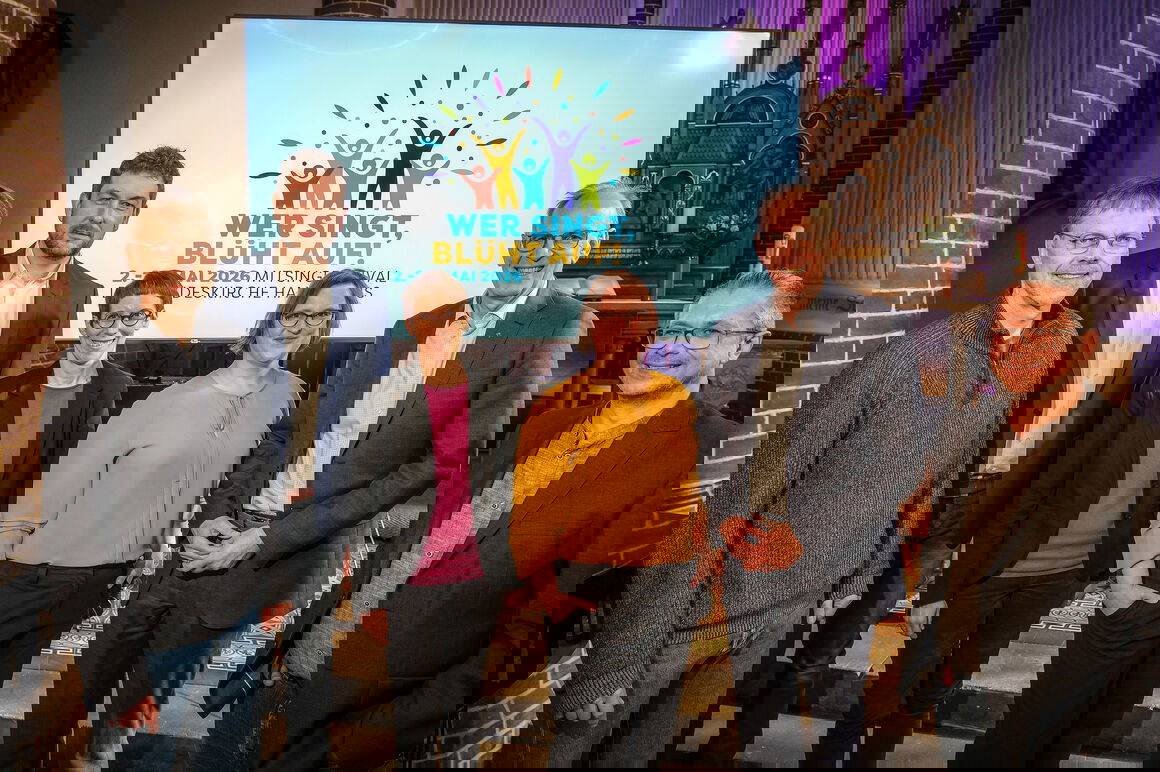 Eine Gruppe von als Männer und Frauen lesbaren Personen steht in einer Kirche vor einer Leinwand mit dem Logo zum Mitsingfestival „Wer singt, blüht auf!“