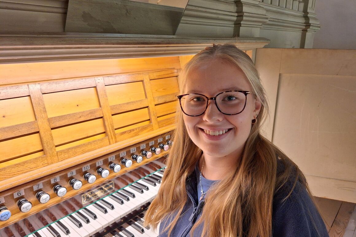 Eine weiblich gelesene, junge Frau mit Brille sitzt an einer Orgel.